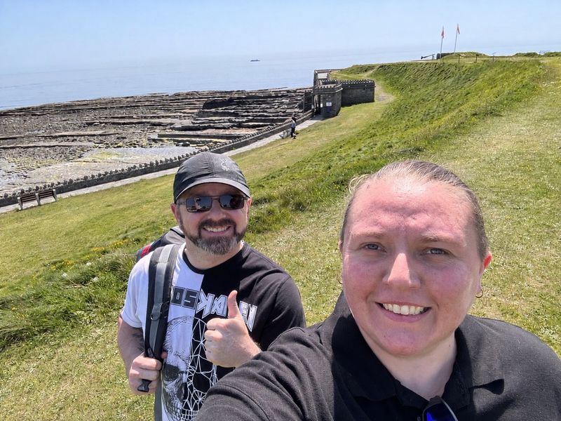 Selfie of Barry Pitts and Samantha Hall smiling on a sunny grassy clifftop on the Isle of Man. Barry, on the left, wears a black cap, sunglasses and a backpack and gives a thumbs up; Samantha is in the foreground in a black top. Behind them are flat rocky shore ledges, a small stone fort and a calm blue sea.