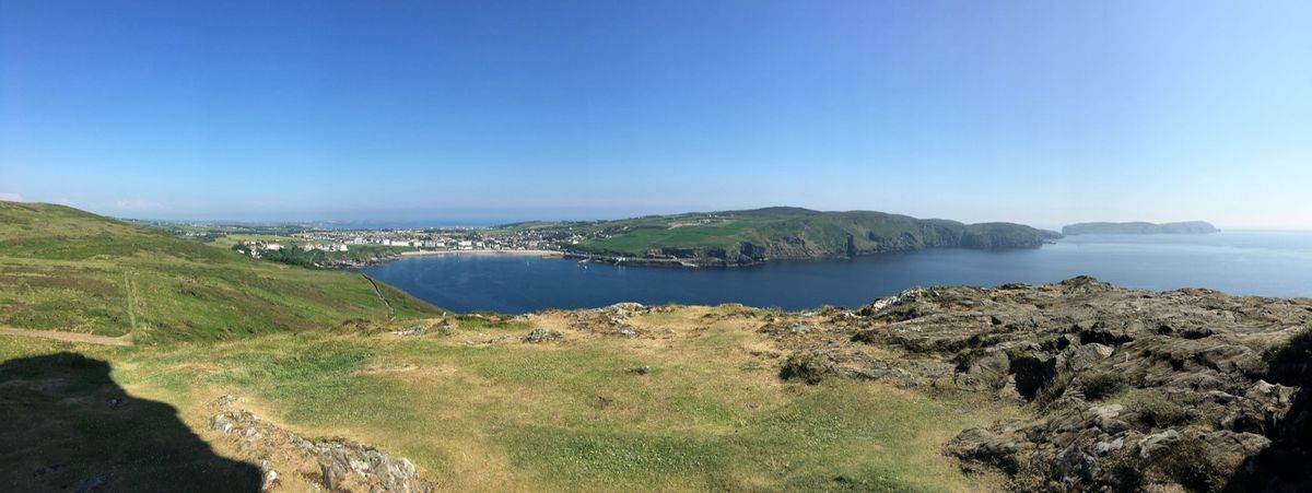 Panoramic view from Bradda Head on the Isle of Man, looking over the deep blue bay of Port Erin to the town, green hills and rugged headlands under a clear blue sky.